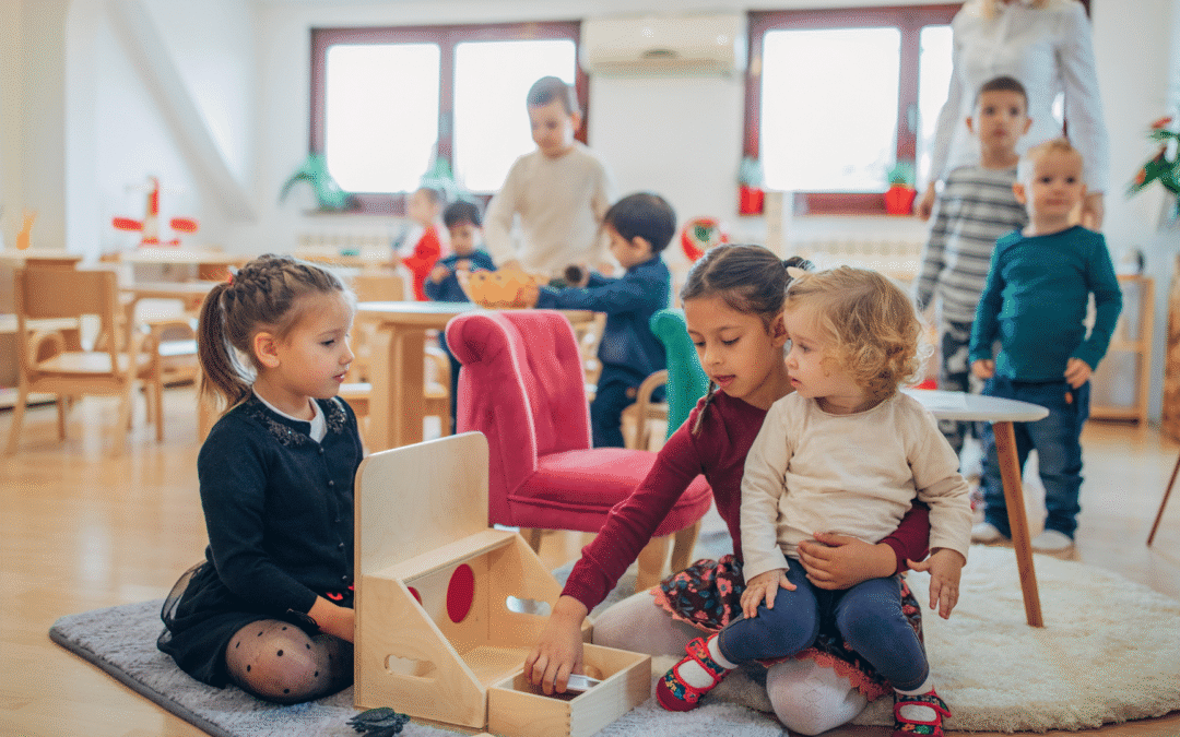 Two young girls sit on a rug and play with a wooden toy house in a bright classroom while other children play in the background central sceneichen