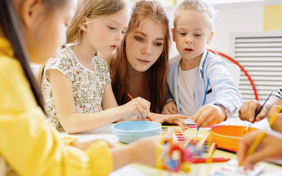 Teacher guiding three children as they paint colorful artwork at a sunny classroom table, cones and bowls visible on the table.
