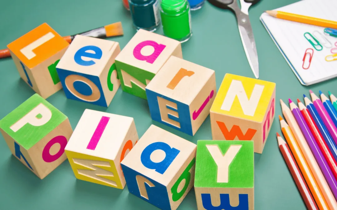 Colorful wooden alphabet blocks scattered on a green desk with scissors, pencils, clips, and a notebook nearby.